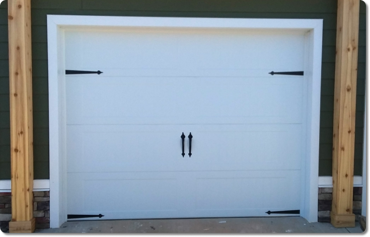 a white garage door with black accents on the front of a forest-green home with wooden beams on either side